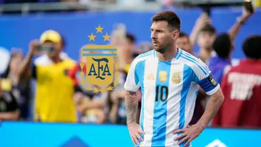 Lionel Messi looks to the side with his hands on his hips while wearing the Argentina national team jersey and the Argentina national team badge is next to him.