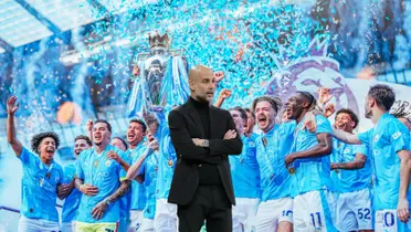 Pep Guardiola crosses his arms looking serious while the Manchester City players celebrate the title with the trophy.