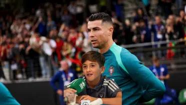 Cristiano Ronaldo looks at the pitch while wearing the Portugal training kit and Thiago Messi is speaking with a microphone.