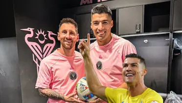 Lionel Messi and Luis Suarez hold a ball together while Cristiano Ronaldo points up with a smile on his face.